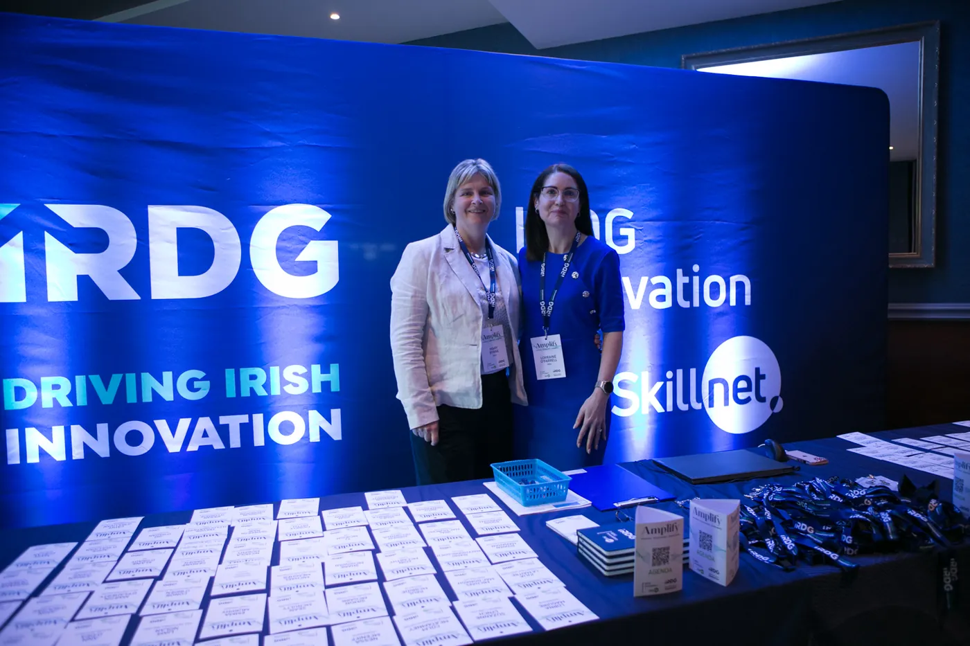 Two members of the IRDG team at the IRDG Innovation Skillnet stand, delegate badges laid out in front of them.